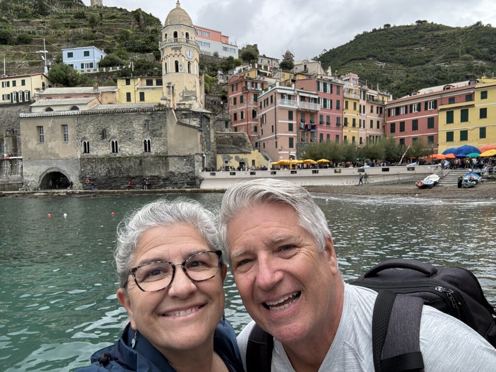 Matt and Linda smile by the waterfront, with colorful buildings behind them and Matt wearing a backpack.